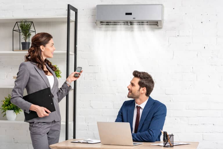 Ein Mann sitzt an einem Schreibtisch mit einem Laptop vor sich und schaut eine Frau an, die in einem grauen Anzug neben ihm steht und eine Fernbedienung in der Hand hält. An der Wand über dem Mann ist eine Klimaanlage zu sehen, aus der Luft strömt. Im Hintergrund befindet sich ein Regal mit Pflanzen.