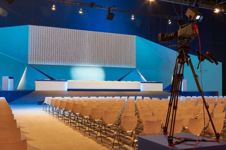 Ein leerer Konferenzraum mit einer großen Bühne im Vordergrund. Eine Kamera auf einem Stativ ist rechts im Bild sichtbar. Die Bestuhlung besteht aus mehreren Reihen von Stühlen. Der Hintergrund ist in Blautönen gehalten.