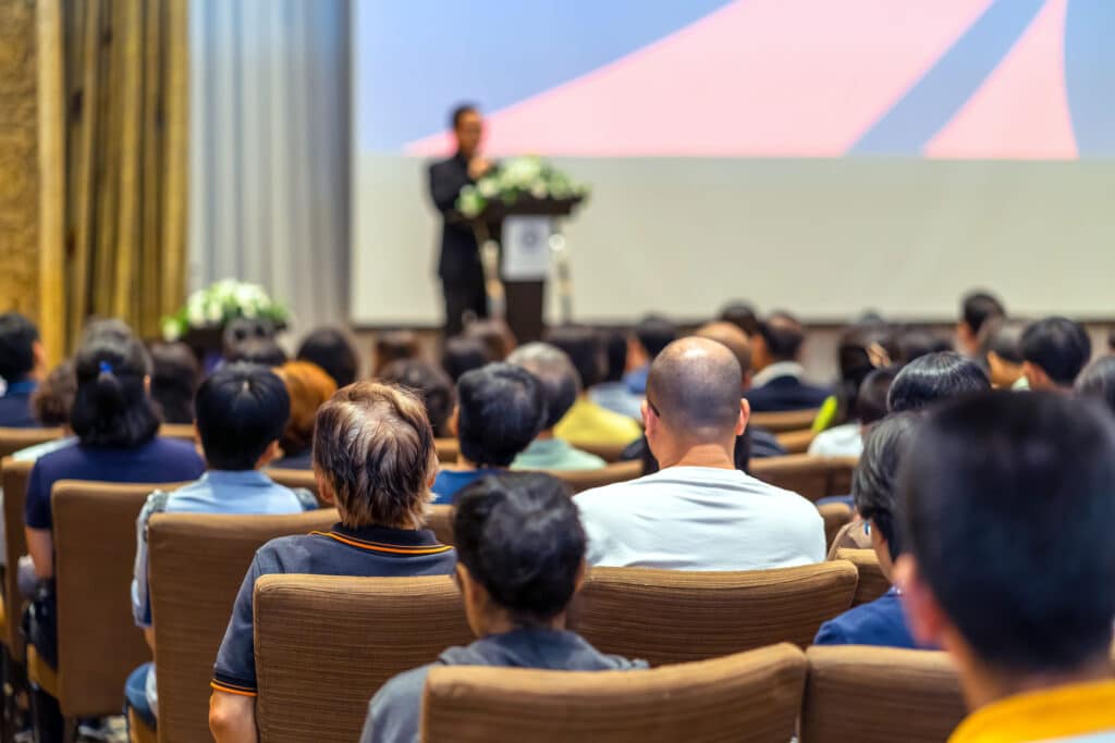 Eine Person steht an einem Rednerpult in einem Konferenzraum, hält eine Präsentation vor einem Publikum von Personen, die auf Stühlen sitzen.