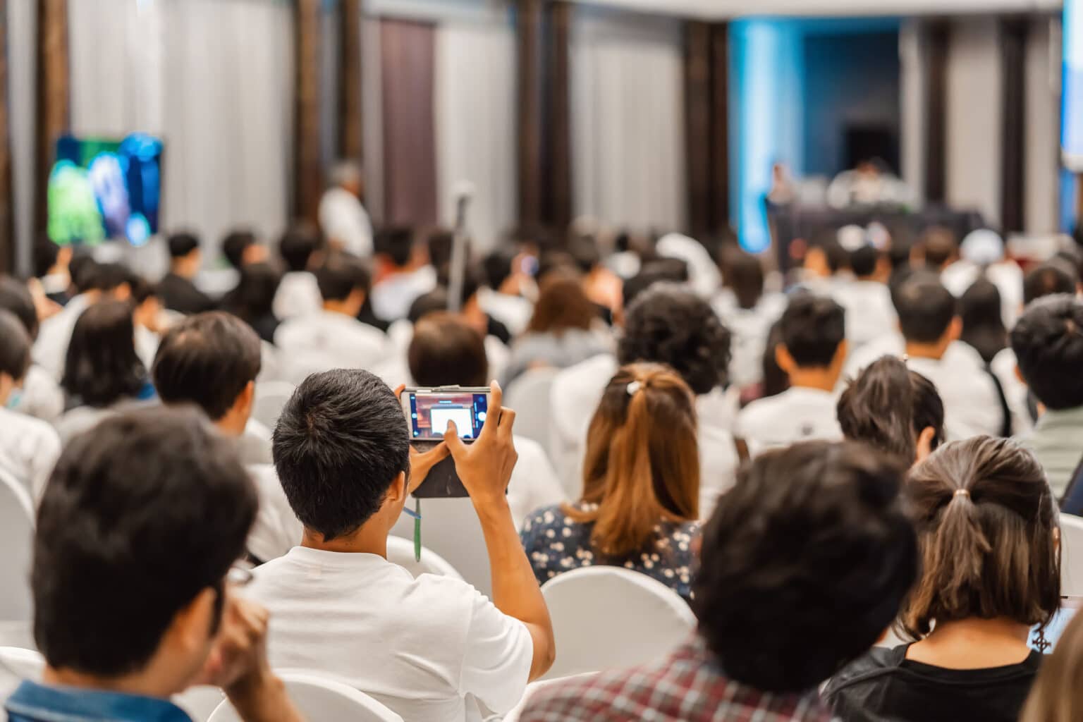 Eine große Gruppe von Menschen sitzt in einem Konferenzraum, einige halten Smartphones hoch, während sie einer Präsentation zuhören.