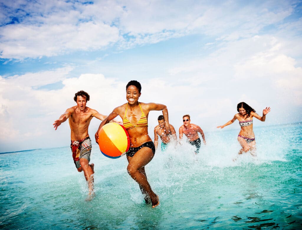 Eine Gruppe von Menschen rennt durch flaches Wasser am Strand. Eine Person vorne trägt einen bunten Strandball. Der Himmel ist teils bewölkt.