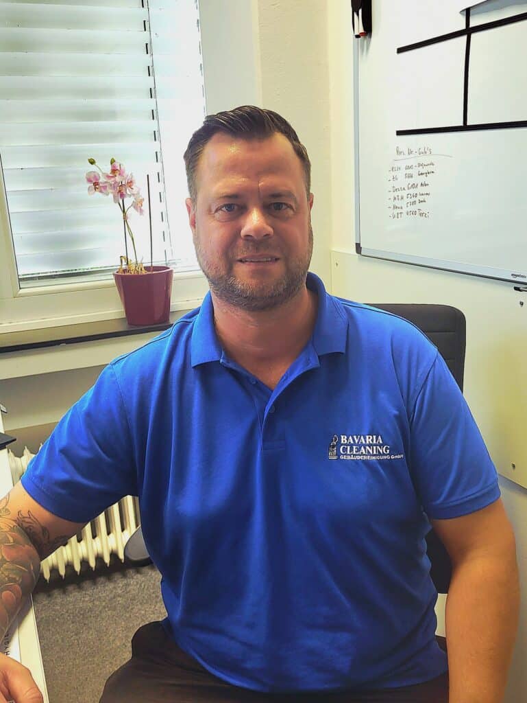 Ein Mann sitzt lächelnd an einem Schreibtisch in einem Büro. Er trägt ein blaues Poloshirt mit dem Logo von Bavaria Cleaning. Im Hintergrund befindet sich ein Fenster mit einer Jalousie und eine weiße Tafel mit Notizen. Auf der Fensterbank steht eine Topfpflanze.
